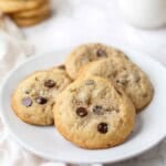 A white plate with browned butter toffee chocolate chip cookies with a glass of milk and extra cookies in the background.