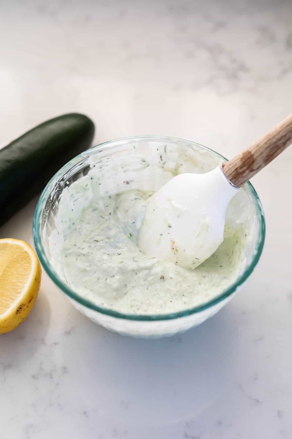 tzatziki sauce in a glass bowl with a rubber white spatula sticking out and a cucumber and lemon wedge on the side