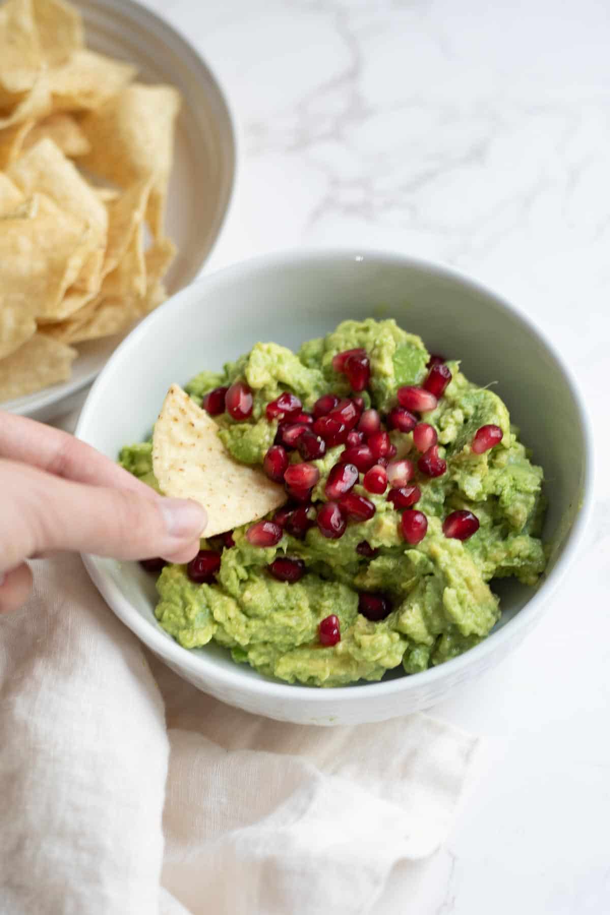 a hand scooping a tortilla chip into a white bowl with guacamole topped with pomegranate seeds