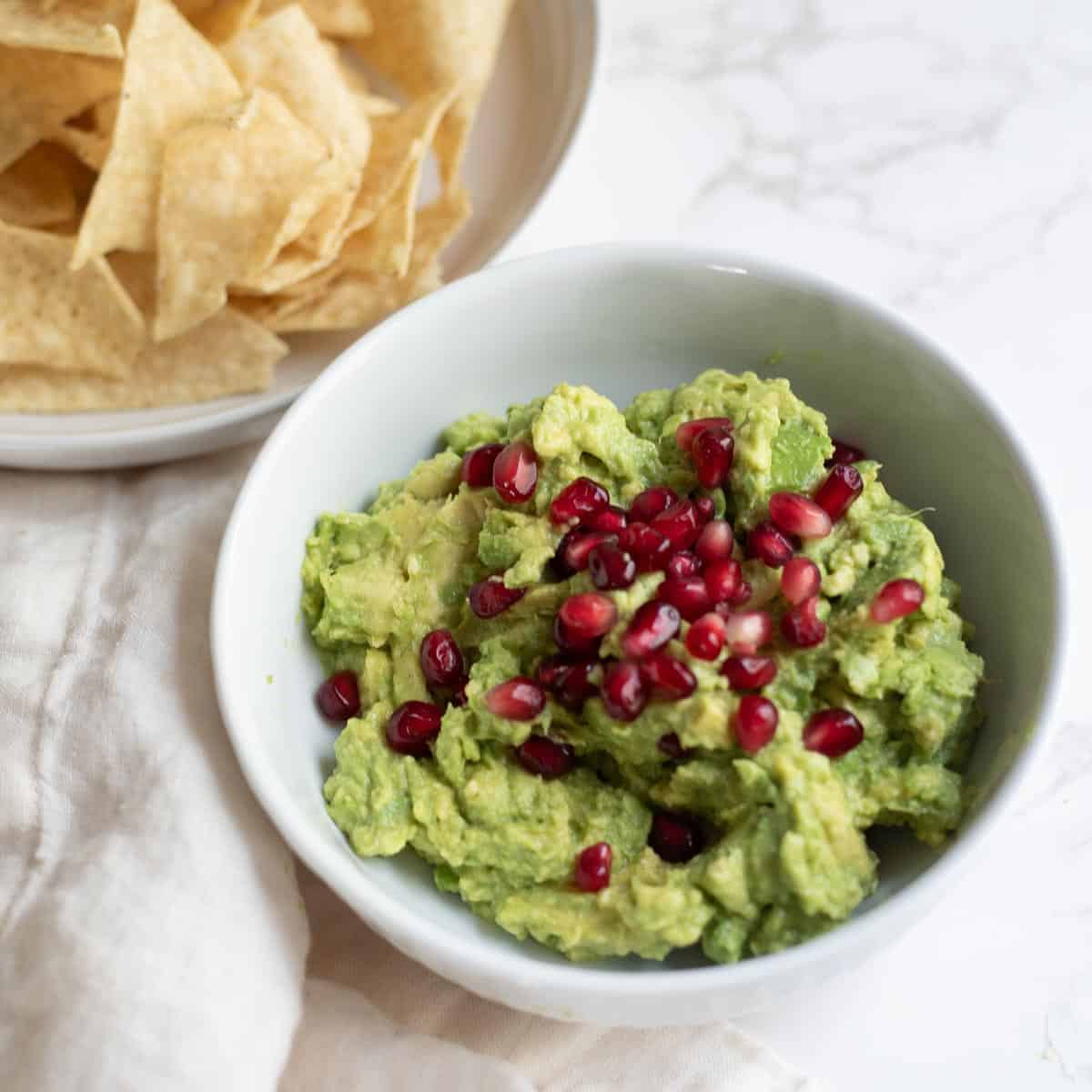 guacamole topped with pomegranate seeds in a white bowl next to a white towel and tortilla chips