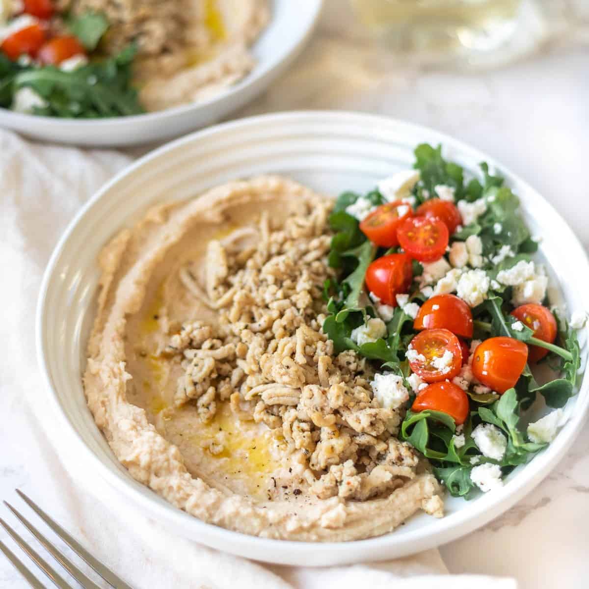 white bowl with white beans, ground chicken, arugula, feta and tomatoes with another plate and glass of white wine in the background