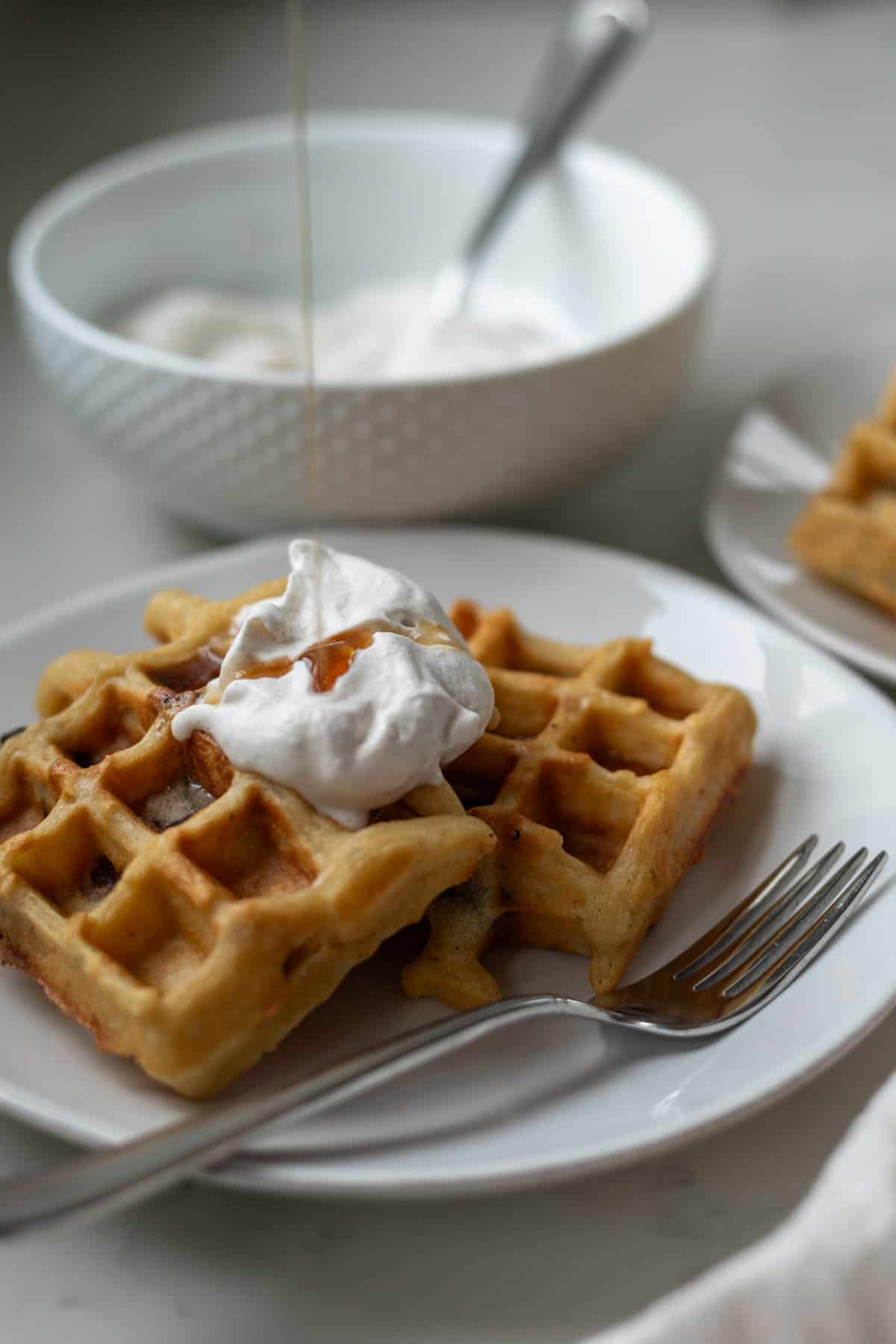 A white plate of waffles with whipped cream on top and maple syrup being drizzled.