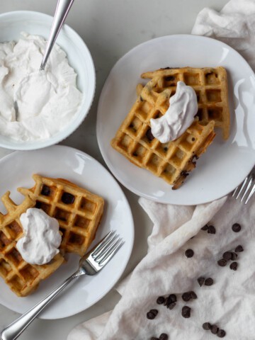 Two white plates of chocolate chip waffles with whipped cream on top.