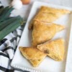 A white plate of Sausage Breakfast Turnovers with Puff Pastry next to a black and white towel, leeks, and eggs.