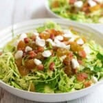 Shaved Brussels Sprout Salad topped with crispy pancetta, raisins, goat cheese, and a warm mustard vinaigrette in a white bowl.