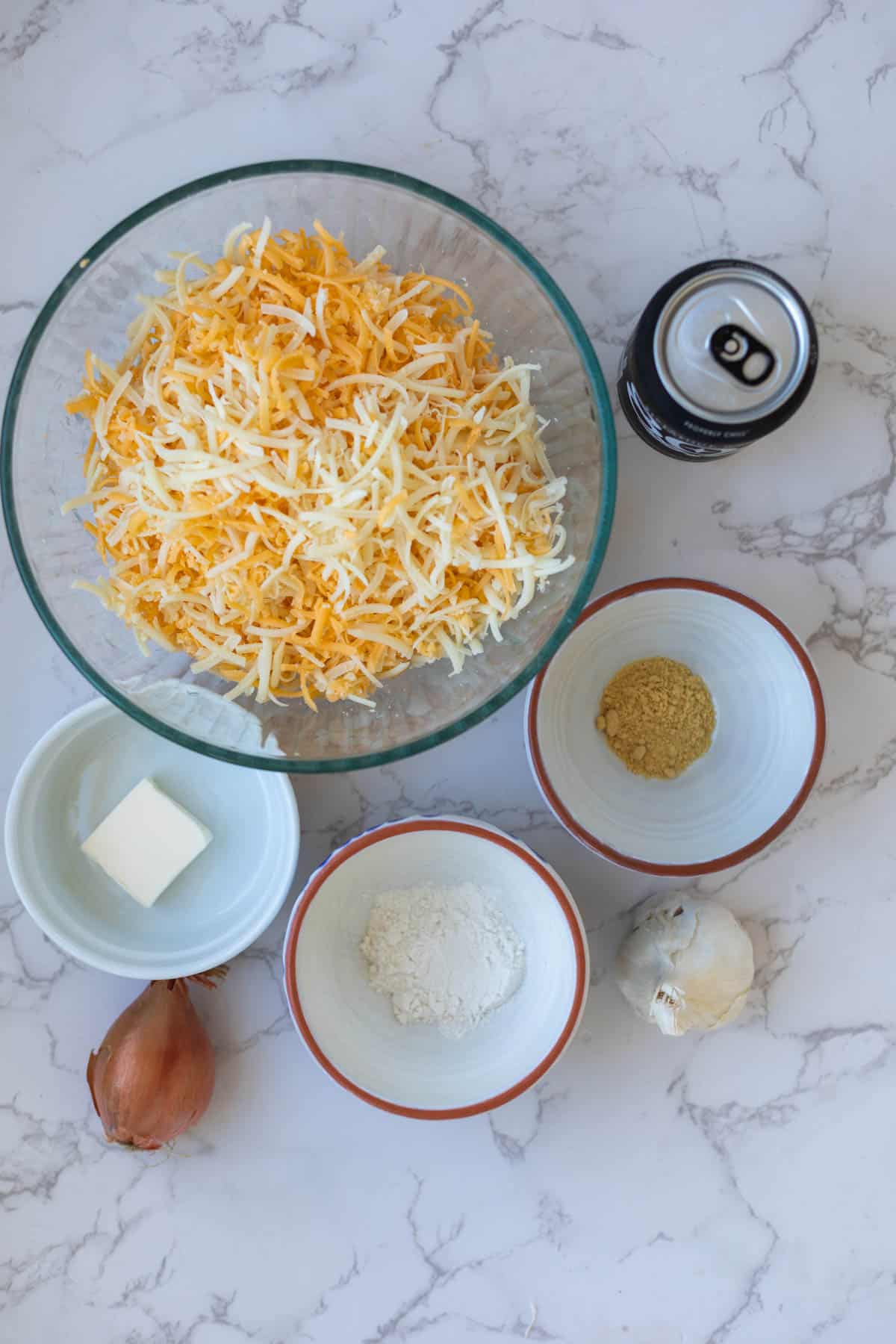 shredded cheese in a bowl next to a can of beer, butter, flour, shallot, garlic, and spices