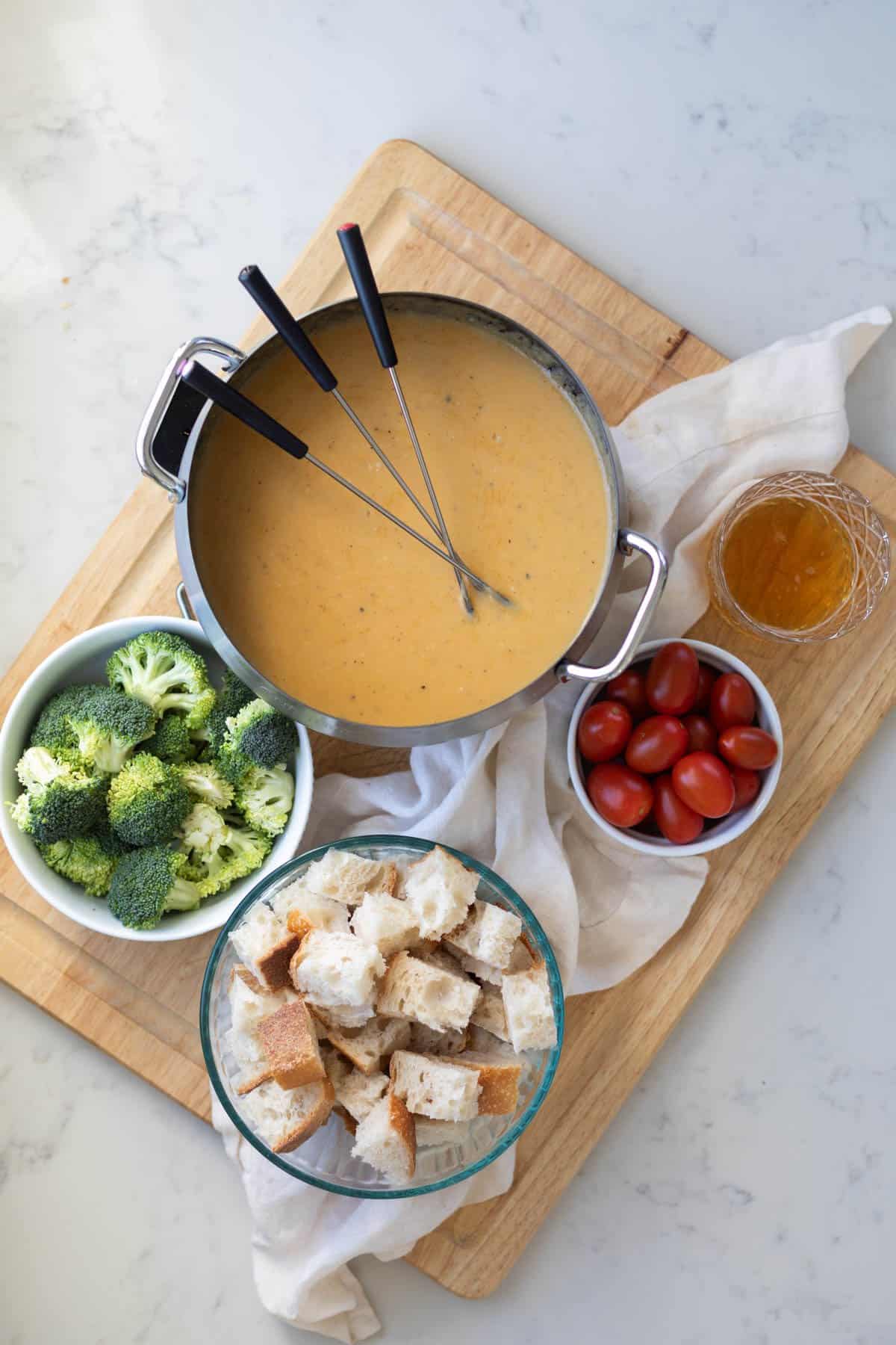 a wooden board with cheese fondue on it along with broccoli, bread cubes, tomatoes, and a glass of beer
