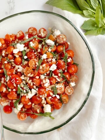 tomato basil feta salad in a clear round bowl with a silver spoon and large basil leaves
