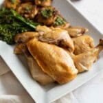 A broken down whole roasted dry brined chicken on a white platter next to potatoes and broccolini.