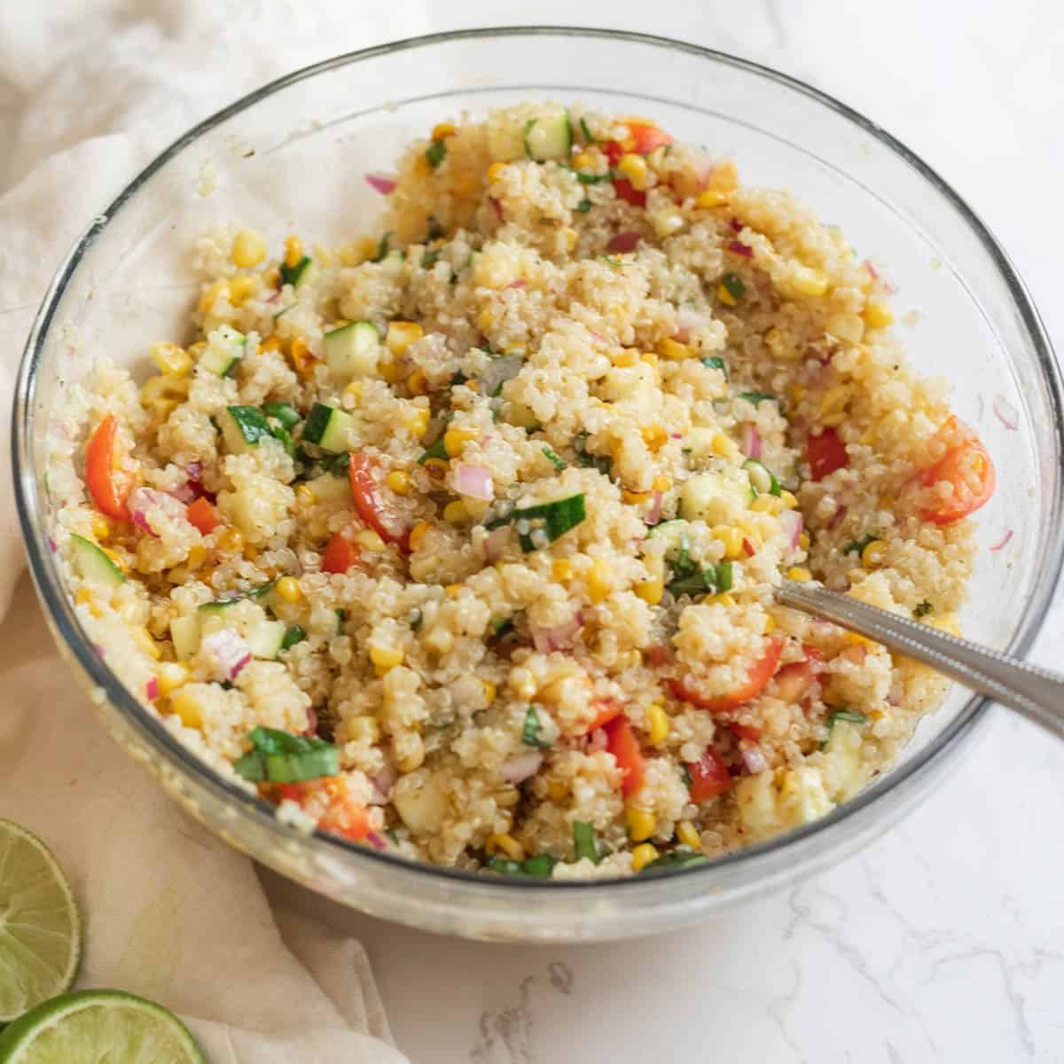 a clear glass bowl filled with quinoa and vegetables with a silver spoon and lime wedges