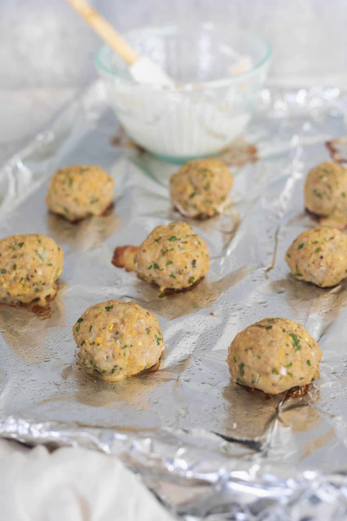 Cooked chicken meatballs on a foil baking sheet with yogurt sauce in the background