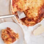An oval pan of Lasanga al Forno with a silver spatula sticking out next to a slice of lasanga on a white plate.