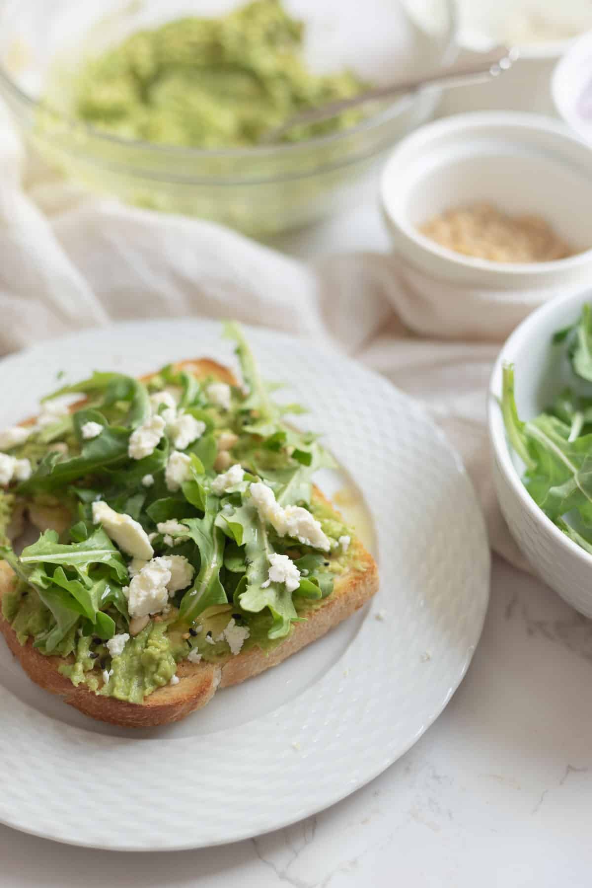 toast on a white plate topped with avocado, arugula, and feta