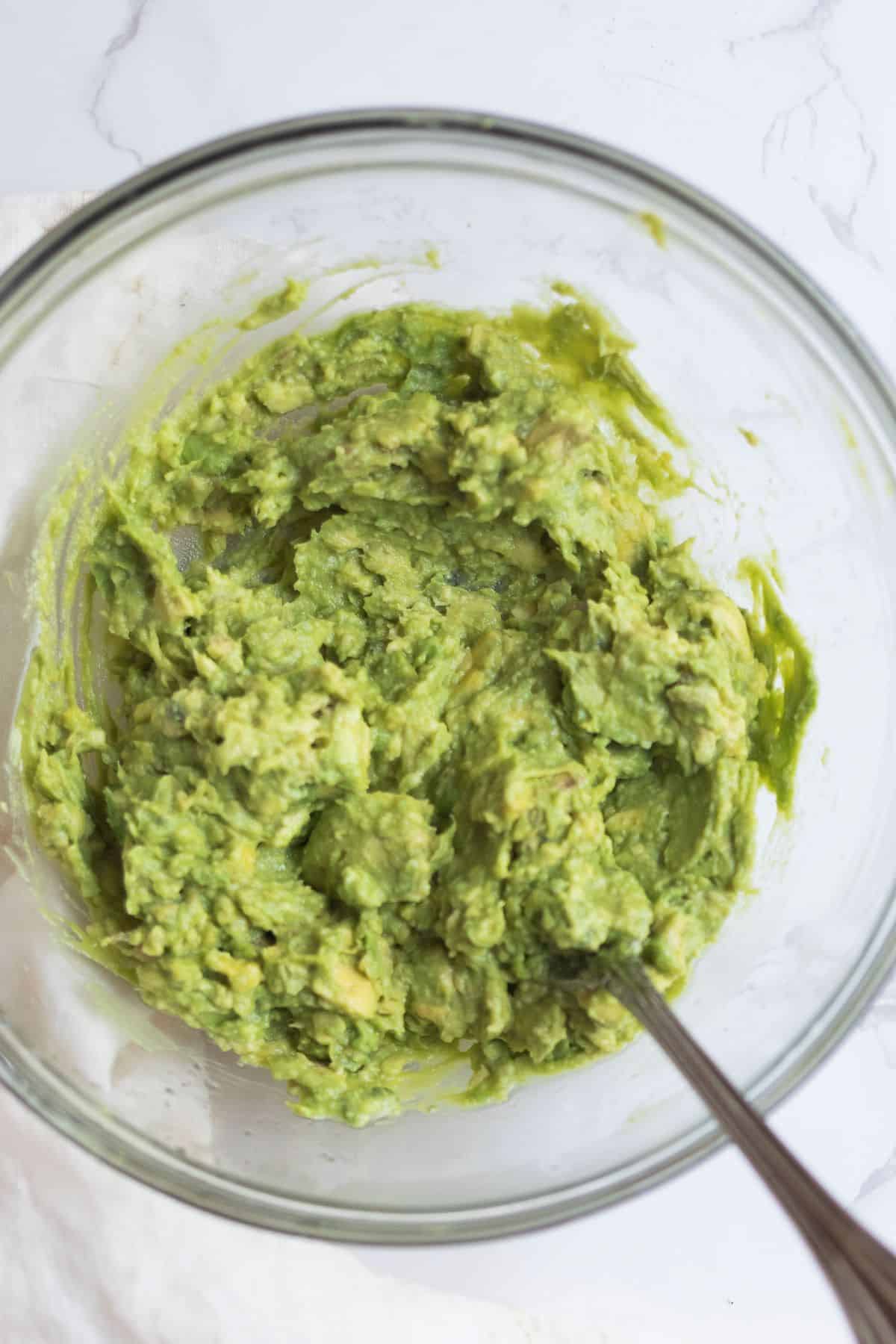 smashed avocado in a glass bowl with a  silver spoon