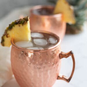 Two copper mugs filled with liquid and ice and garnished with a pineapple wedge