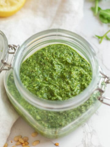 green pesto sauce in a clear jar on a white counter with pine nuts, a lemon half, and arugula in the background