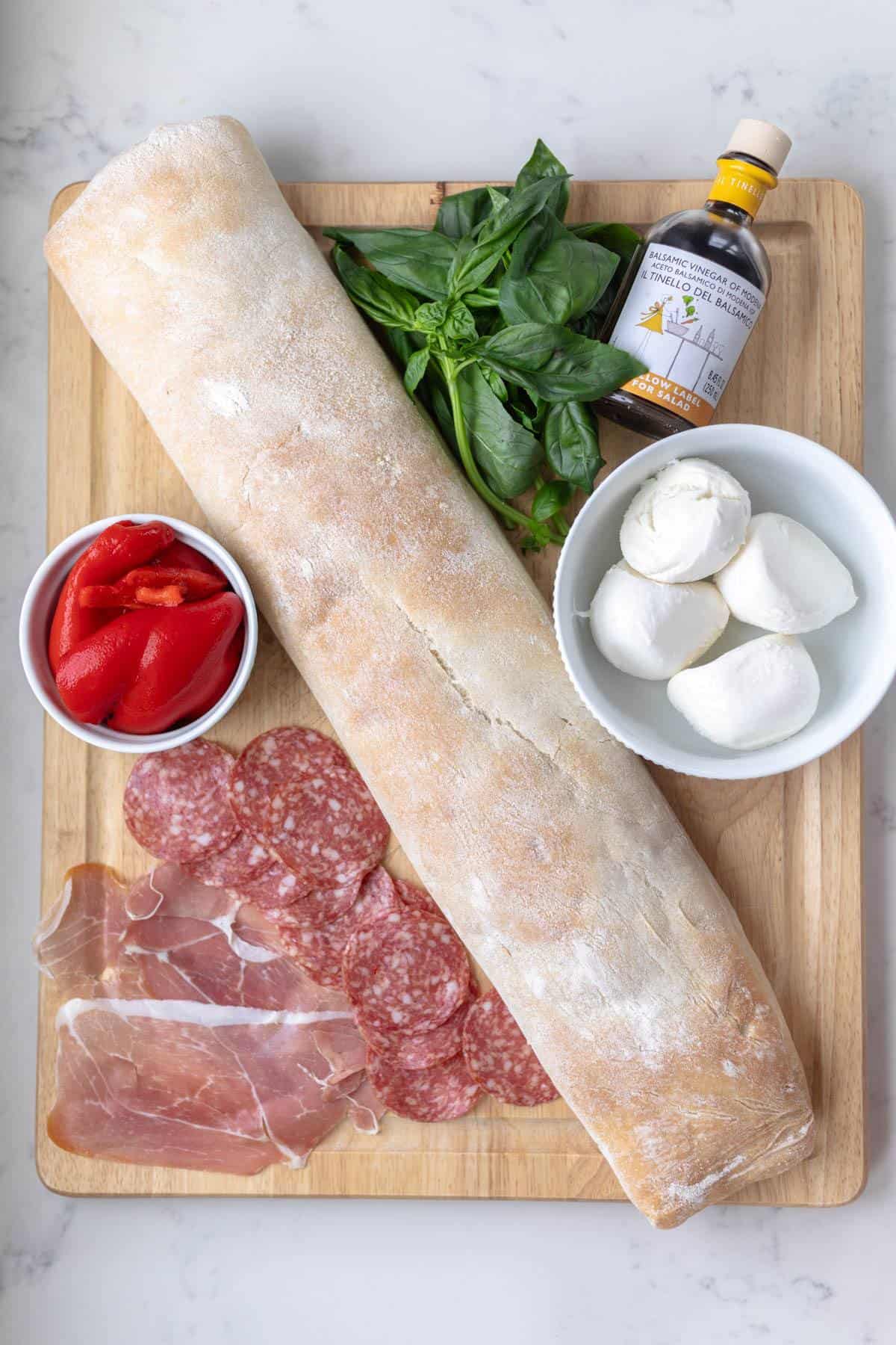 baguette, basil, peppers, balsamic, mozzarella, salami, and prosciutto on a wooden board on a marble counter