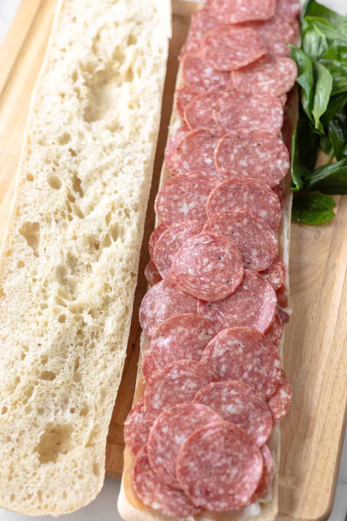 salami layered on a loaf of bread next to plain bread and basil leaves