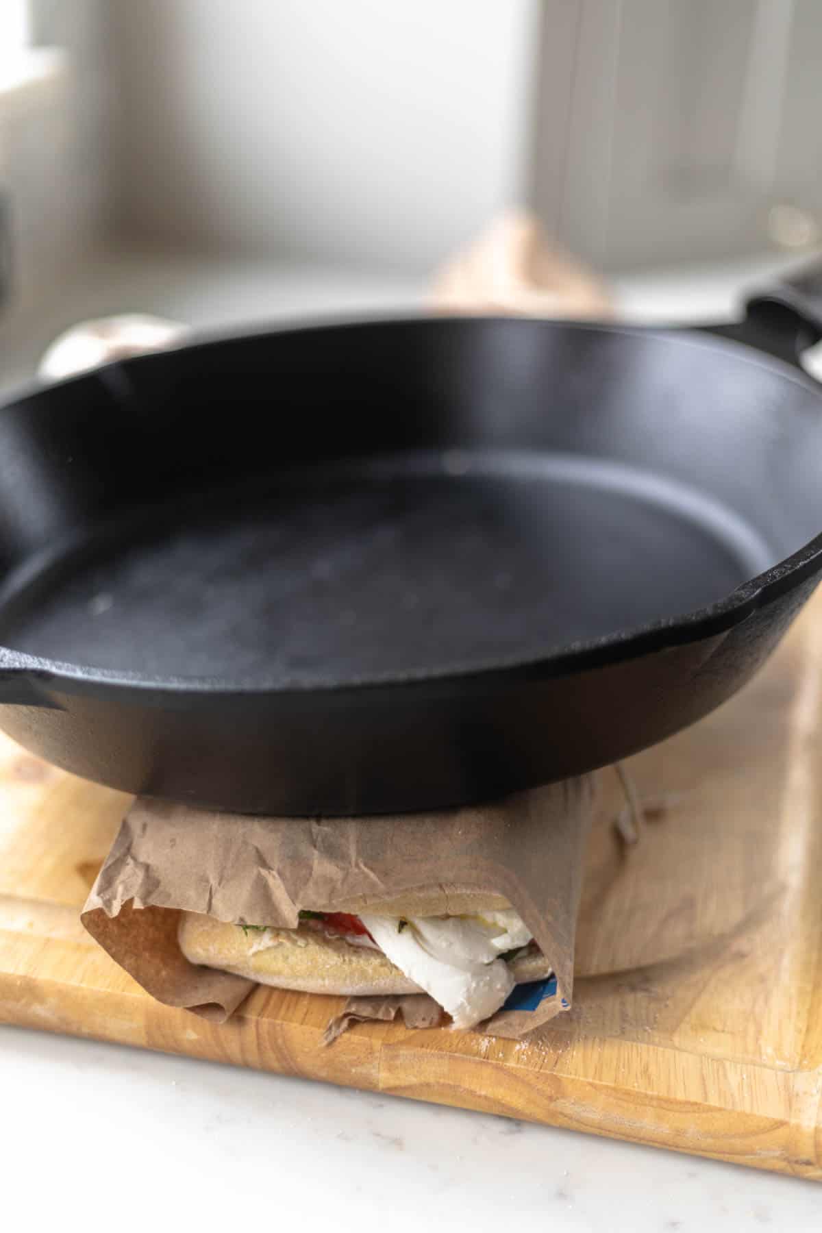 a sandwich on a wooden board being pressed down by a cast iron skillet