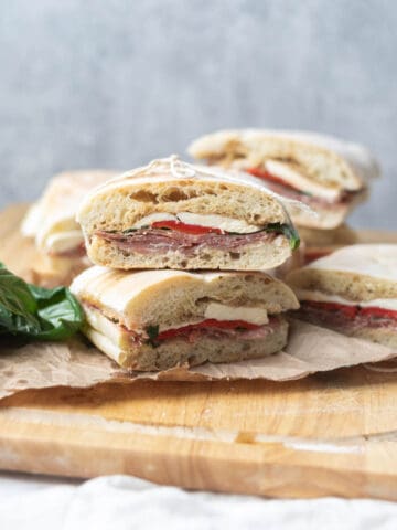 Italian sandwiches stacked on top of each other on a wooden cutting board and brown paper next to basil leaves