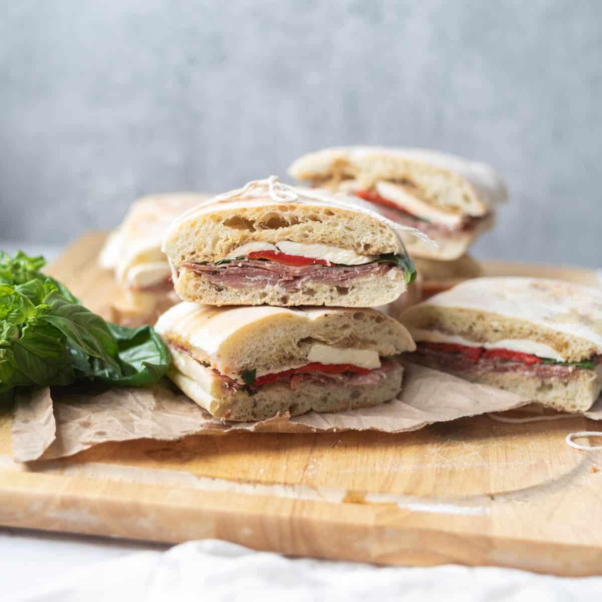 Italian sandwiches stacked on top of each other on a wooden cutting board and brown paper next to basil leaves