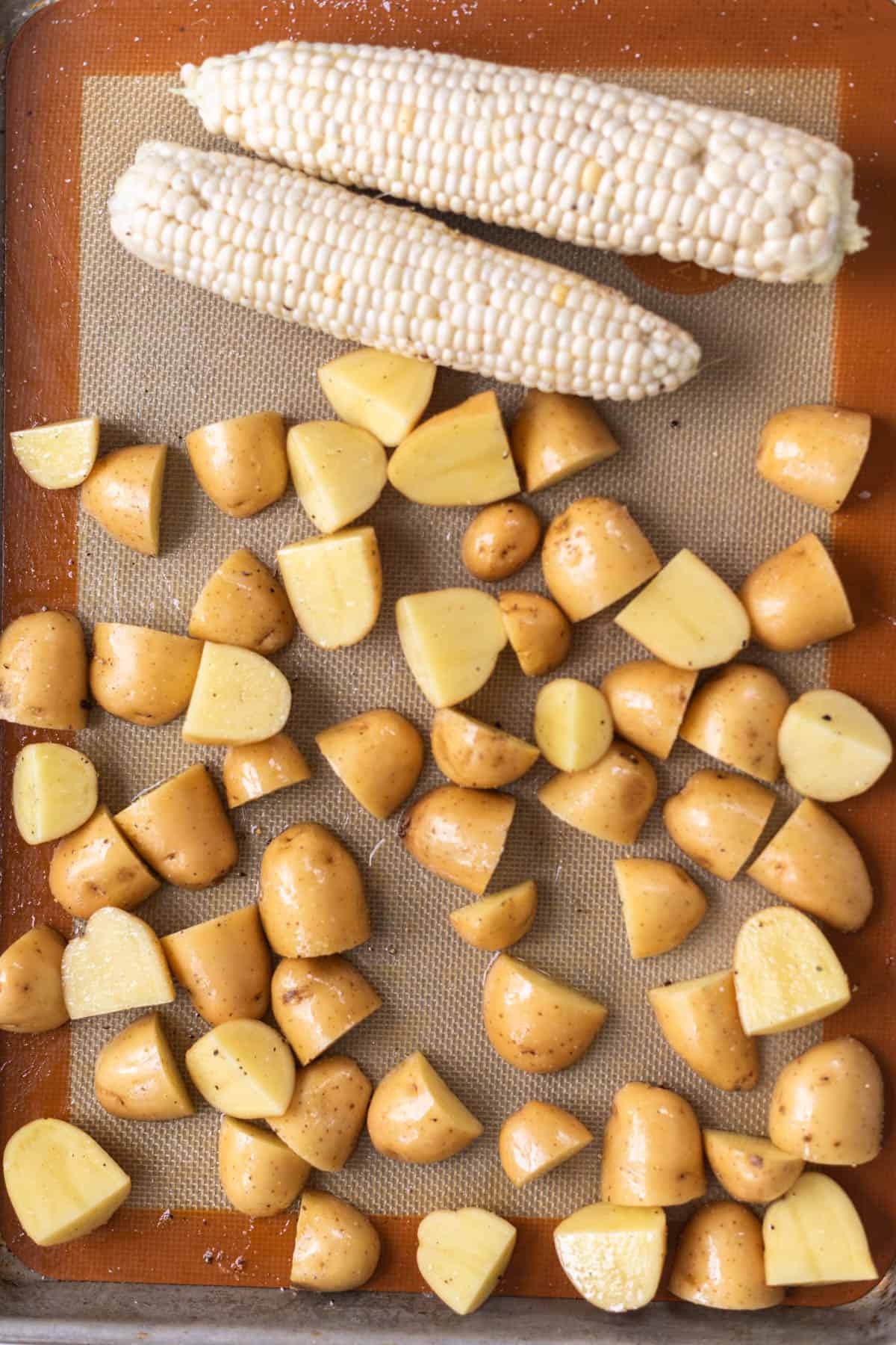 raw quartered yellow potatoes and two corn on the cobs on a baking sheet