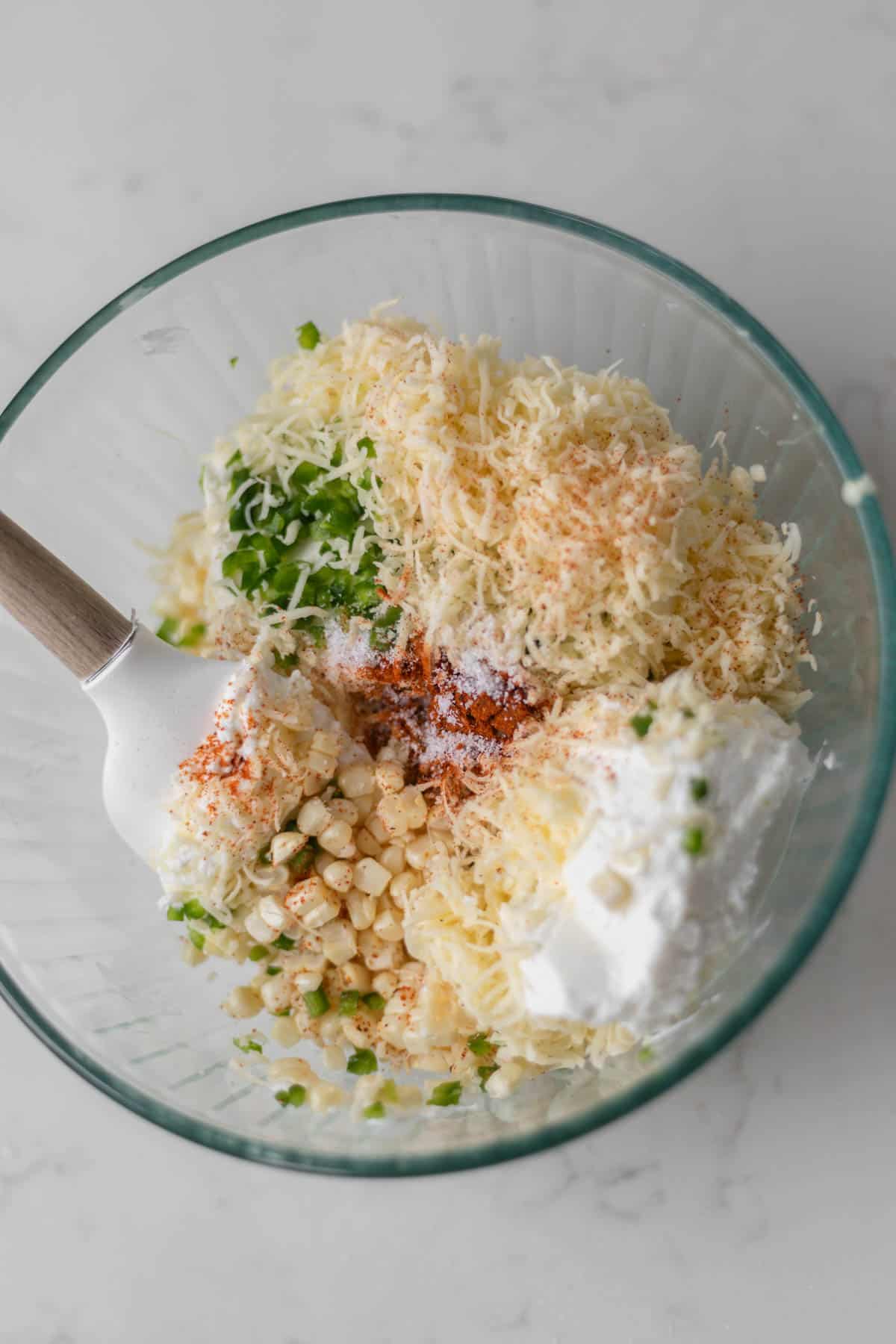 cheese, cream cheese, corn, jalapeno, greek yogurt, and chili powder in a glass bowl with a wooden spoon