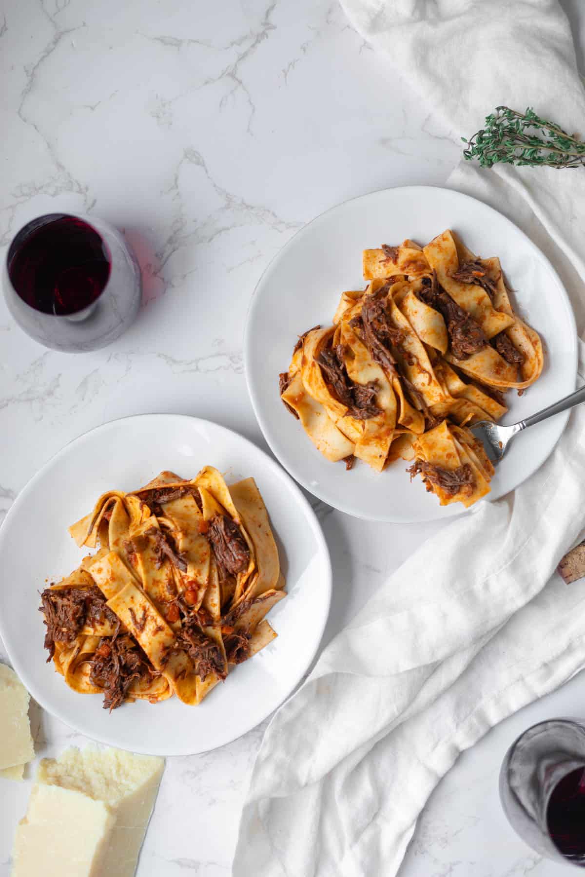 two white plates of short rib ragu next to parmesan, two red wine glasses, and thyme on a white counter