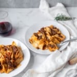 two white plates of pappardelle pasta with short rib ragu next to thyme and a glass of wine on a white napkin