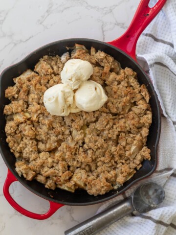 Apple crisp in a red cast iron skillet topped with vanilla ice cream next to a white towel