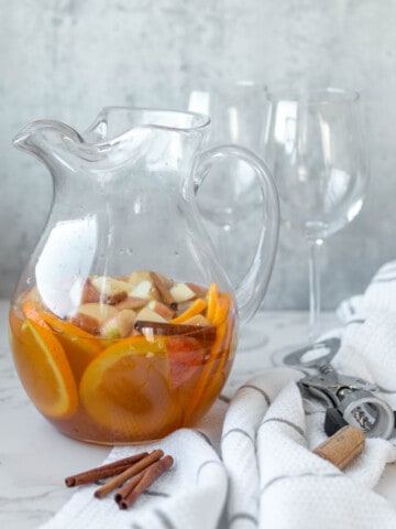 fall sangria in a clear pitcher next to cinnamon sticks, a wine opener, white towel and two glasses
