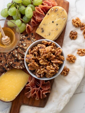 honey walnuts in a bowl on a wooden board next to cheeses, grapes, honey, crackers, salami, and more walnuts
