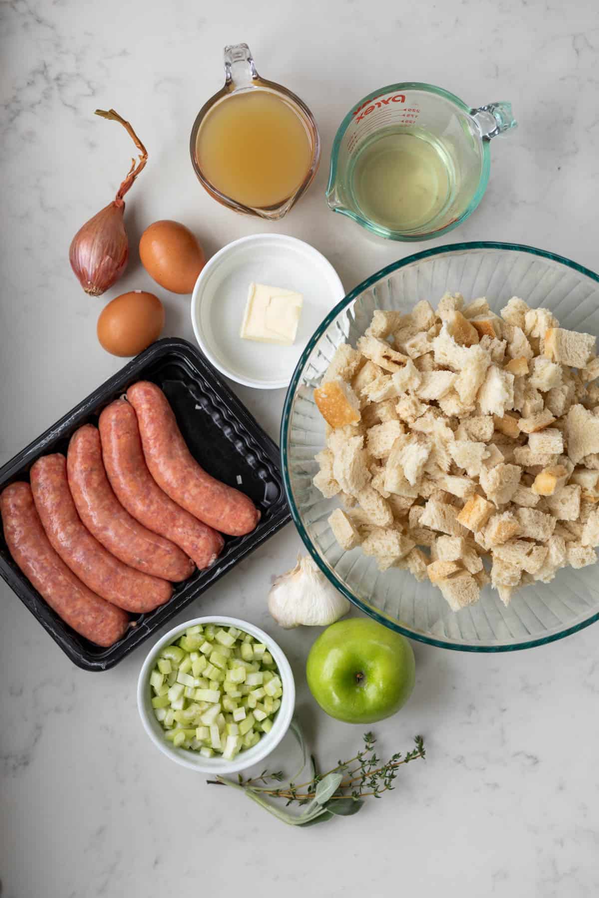 bread cubes in a bowl next to sausage, eggs, butter, shallot, wine, stock, garlic, apple, celery, and herbs