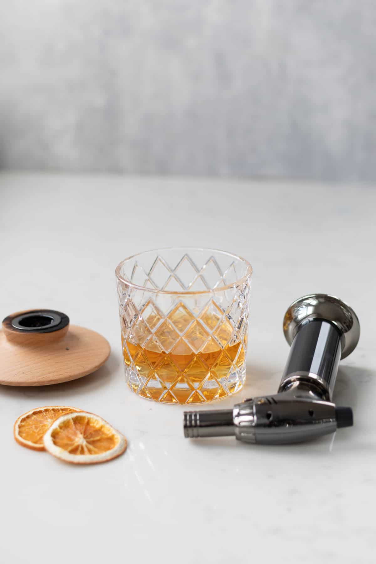 whisky in a glass next to two orange slices and a cocktail smoker