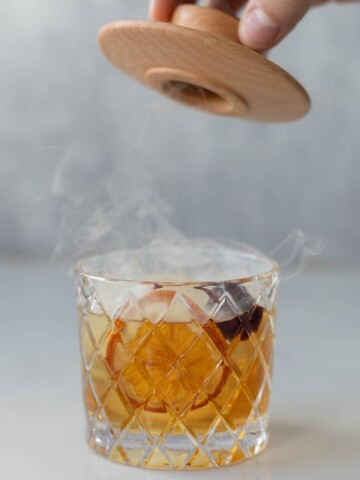 an old fashioned in a lowball glass with smoke coming out of it and a hand lifting a lid over it