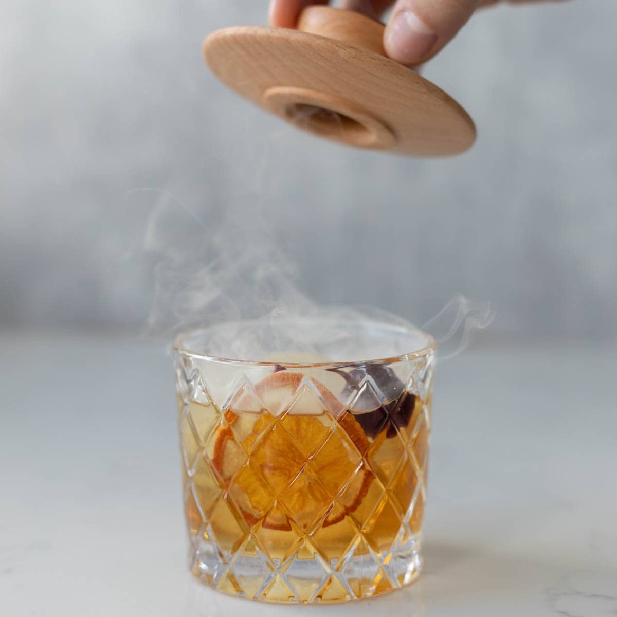 an old fashioned in a lowball glass with smoke coming out of it and a hand lifting a lid over it