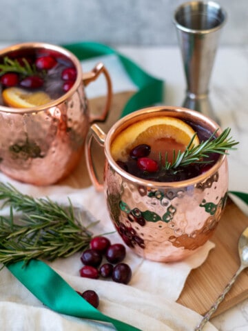 two christmas moscow mules in copper mugs next garnished with rosemary, cranberries, and orange next to a green ribbon