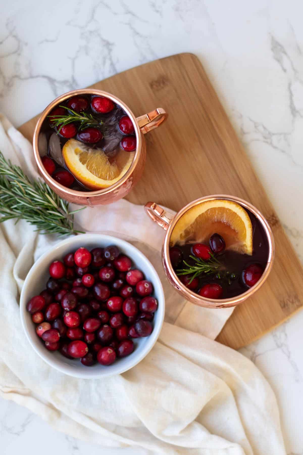an overhead photo of two christmas mules in a copper mug on a wooden cutting board next to cranberry and rosemary garnishes