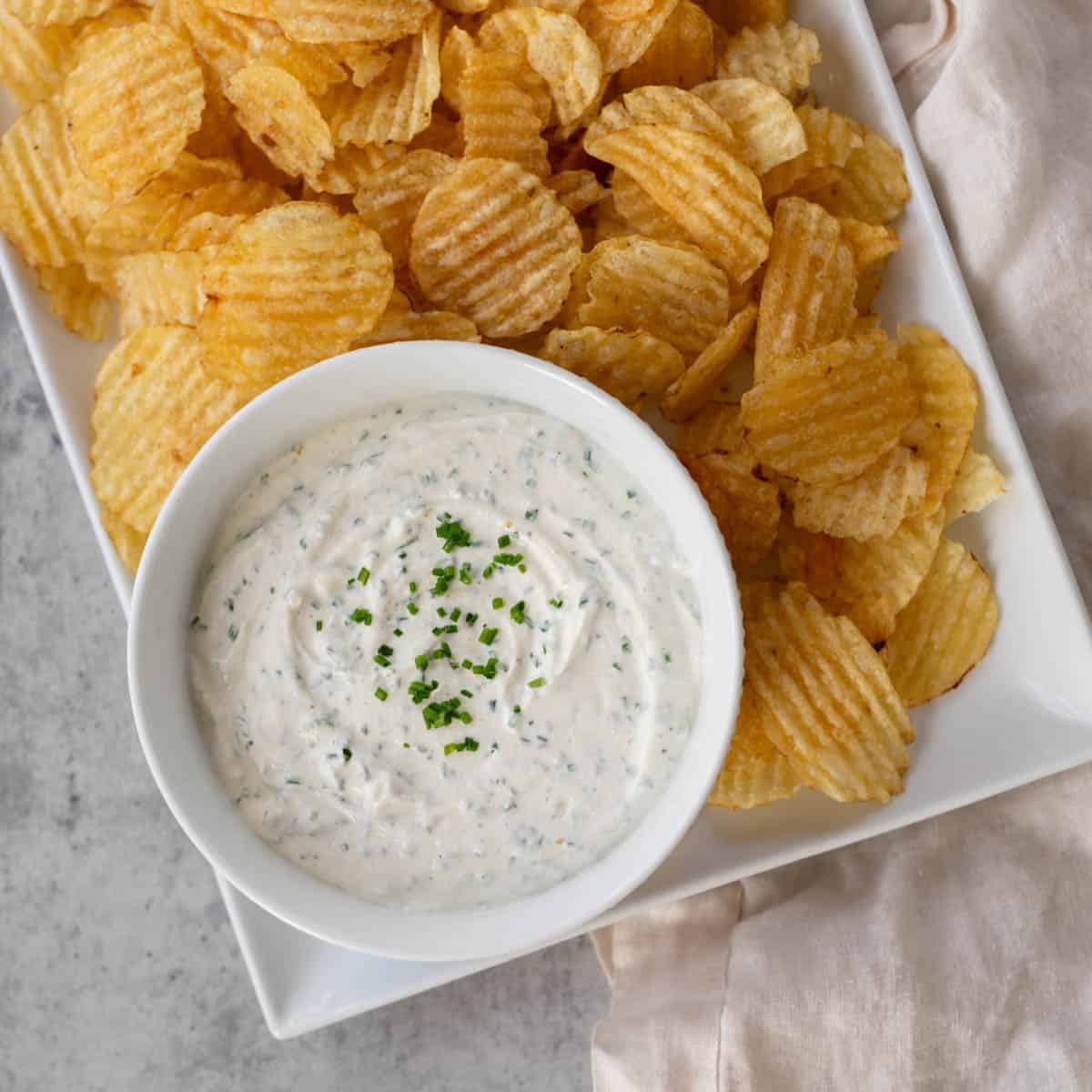 sour cream and chive dip in a white bowl on a rectangular platter filled with potato chips