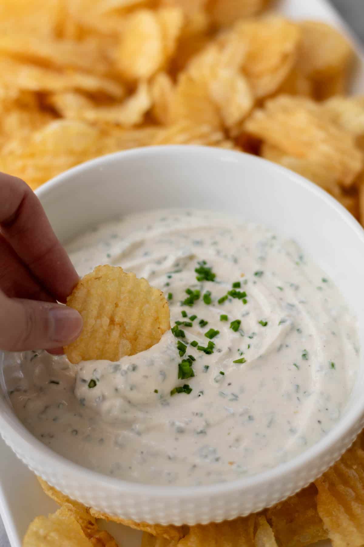 a hand dipping a potato chip into sour cream dip in a white bowl