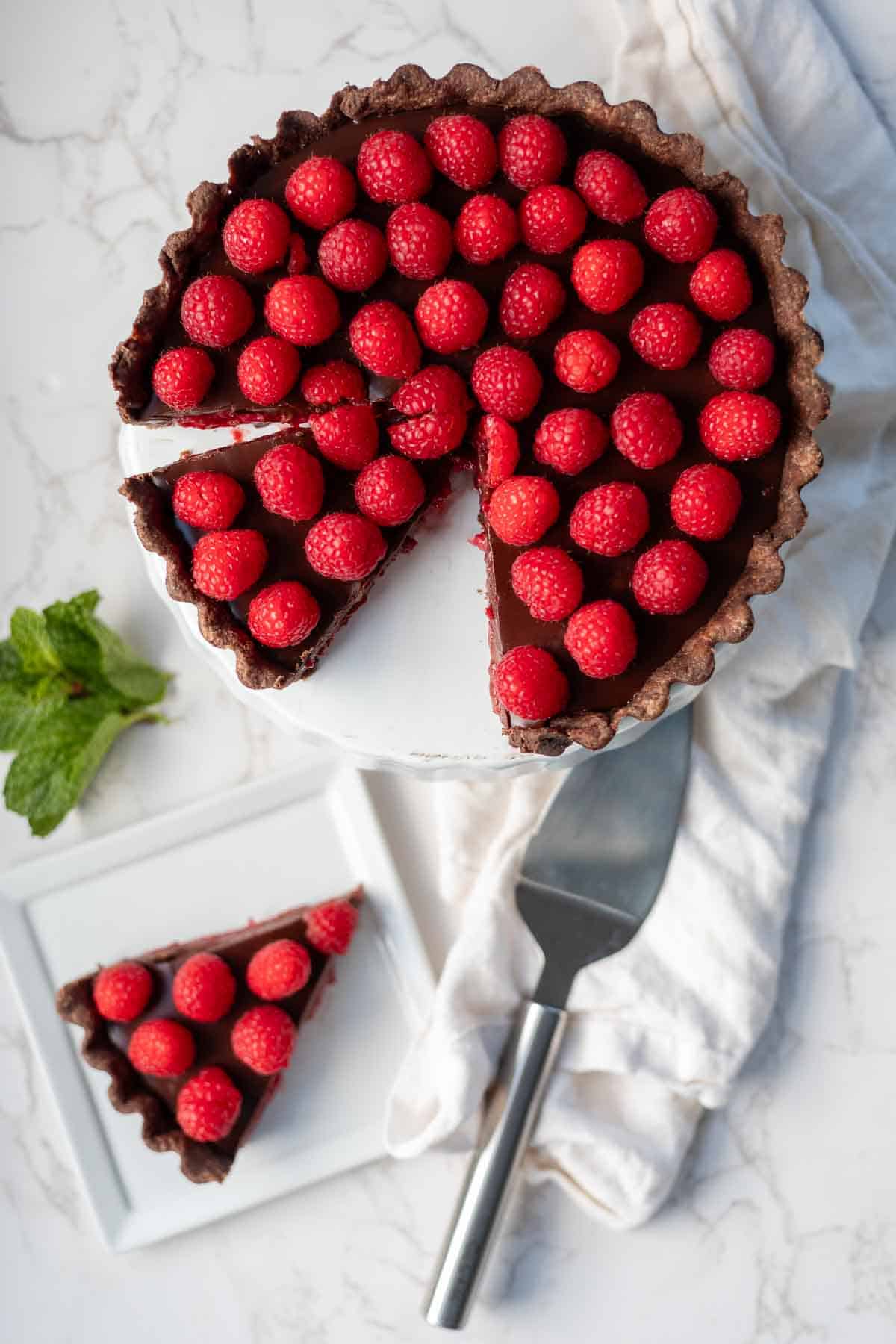 Chocolate raspberry tart on a pie dish next to a slice of tart, mint, and a spatula.