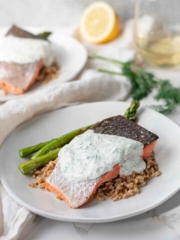 Two plates of salmon with a yogurt dill sauce on top of farro and asparagus next to a glass of wine.