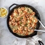 Shrimp scampi and angel hair pasta in a cast iron skillet next to lemon wedges on a white counter.