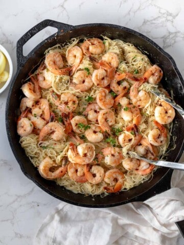 Shrimp scampi and angel hair pasta in a cast iron skillet next to lemon wedges on a white counter.