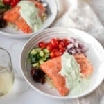 Two plates of salmon on top of Greek quinoa salad and topped with tzatziki sauce next to a glass of white wine.