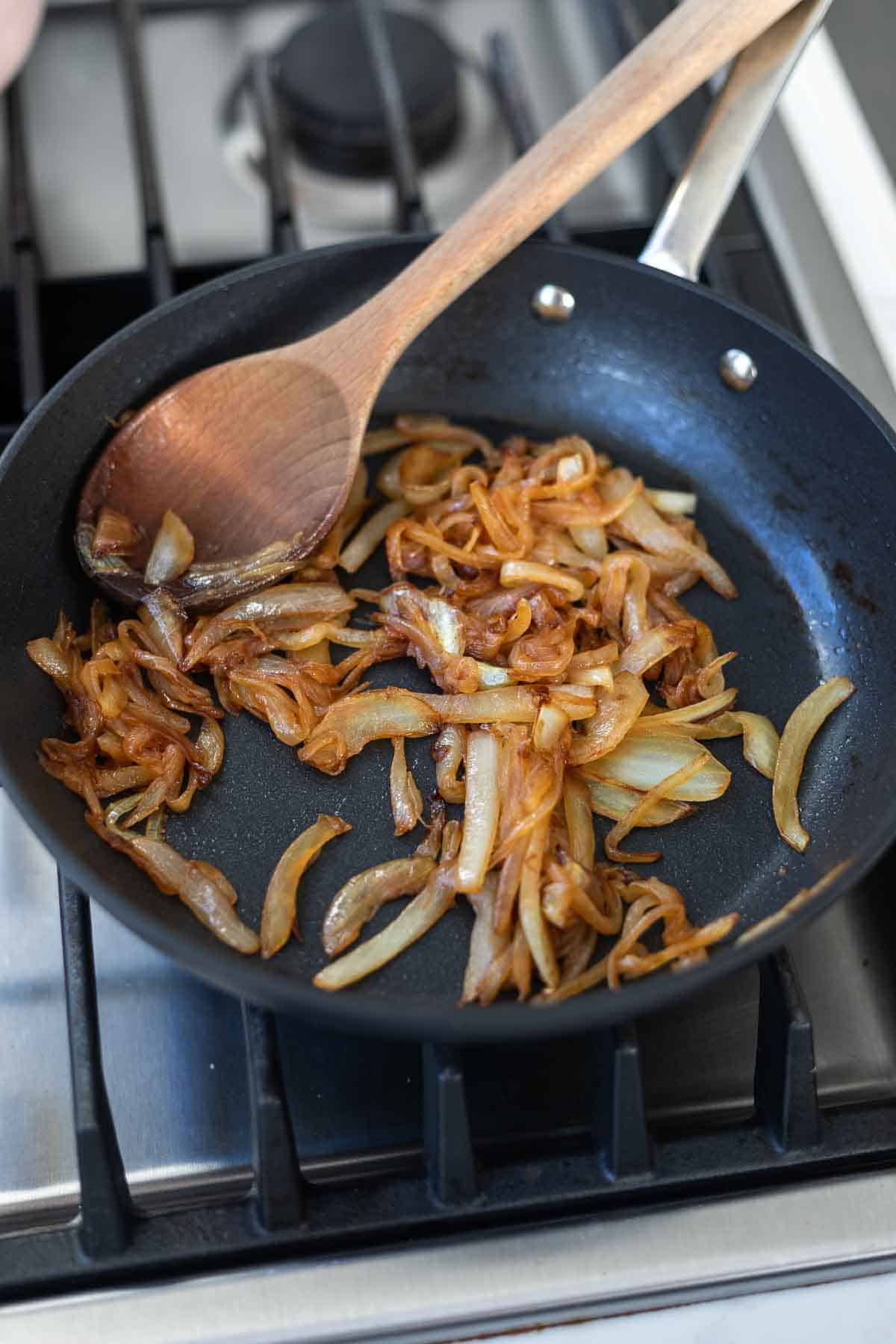 Sliced caramelized onions in a black pan on the stove with a wooden spoon.