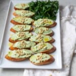 Platter of green garlic herb whipped goat cheese crostini topped with pine nuts next to fresh herbs.