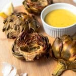 Roasted artichokes on a cutting board with garlic and homemade aioli in a white bowl.