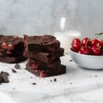 Fudgy chocolate cherry brownies stacked next to a bowl of fresh cherries, chocolate chips, and a glass of milk.