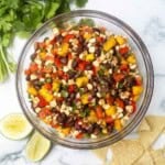 A bowl of black bean and mango salsa on a white counter next to cilantro, tortilla chips, and lime wedges.